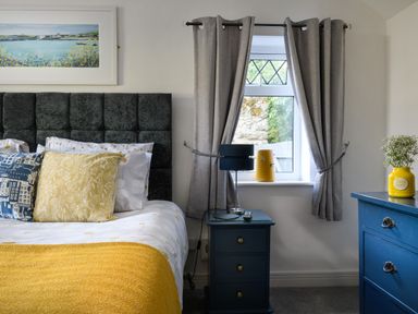 A bedroom with a bed with patterned pillows a yellow blanket blue bedside table with a lamp and window with grey curtains at Ty Bach - Beaumaris in Beaumaris