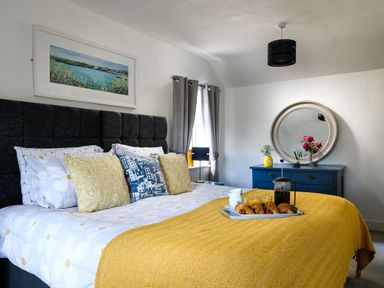 A bedroom with a bed covered in pillows and a yellow blanket with a tray of croissants and coffee on it and a blue dresser with a round mirror above it at Ty Bach - Beaumaris in Beaumaris