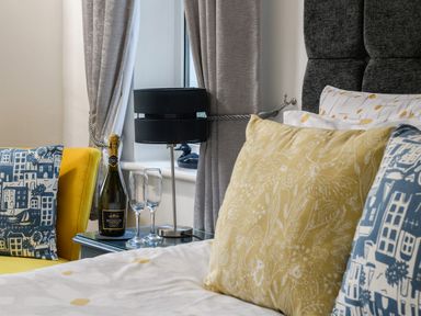 A bedroom corner with a bed featuring decorative pillows next to a yellow chair and a table with a bottle of sparkling wine and two glasses at Ty Bach - Beaumaris in Beaumaris