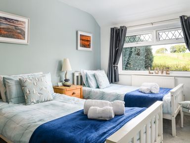 A bedroom with two single beds with blue blankets and seagull cushions a wooden nightstand with a lamp between the beds and a large window with dark curtains at Ty Bach - Beaumaris in Beaumaris