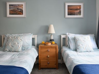 A bedroom with two single beds, a wooden nightstand with a lamp, croissants, and orange juice between the beds at Ty Bach - Beaumaris