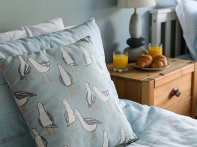 A bed with a pillow printed with seagulls and a wooden nightstand with a lamp croissants and two glasses of orange juice at Ty Bach - Beaumaris in Beaumaris