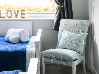 A bedroom corner with a chair featuring bird print cushions and a bed with rolled white towels on a blue blanket at Ty Bach - Beaumaris in Beaumaris