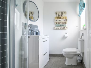 A bathroom with a shower, white vanity with mirror, toilet, wall decoration, and window at Ty Bach - Beaumaris in Beaumaris