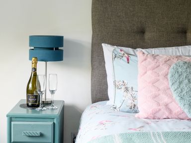 A bedside table with a blue lamp bottle of Nozeco sparkling wine and two empty glasses next to a bed with floral sheets and pink and green pillows at Ty Bach - Beaumaris in Beaumaris