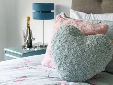A bedroom with patterned bedding heart shaped pillows a blue lamp and a bottle with two glasses on a bedside table at Ty Bach - Beaumaris