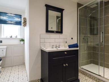 A bathroom with a black vanity cabinet and mirror a shower with glass door and tiled walls a toilet under a window with a blue curtain and patterned floor tiles at Ty Bach - Beaumaris in Beaumaris