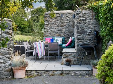 An outdoor seating area with a stone wall a table with chairs cushions and a barbecue at Ty Bach - Beaumaris in Beaumaris