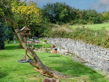 A garden with a tree trunk a bird feeder stone wall and two metal chairs with pink cushions at Ty Bach Beaumaris