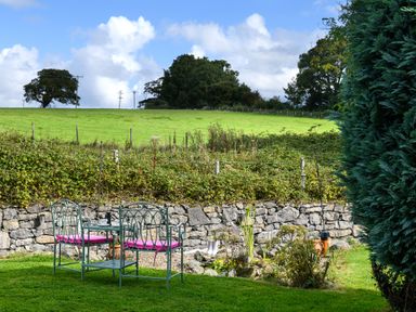 A garden with metal chairs and table with pink cushions near a stone wall and green fields at Ty Bach - Beaumaris in Beaumaris