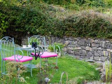 A garden area with two metal chairs with pink cushions and a small table with cups and binoculars by a stone wall at Ty Bach - Beaumaris in Beaumaris