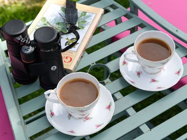 Two cups of coffee in floral china cups on a green slatted table with binoculars and a book at Ty Bach - Beaumaris