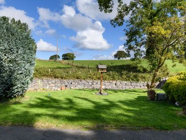 A garden with a bird feeder on grass a stone wall and trees at Ty Bach - Beaumaris in Beaumaris