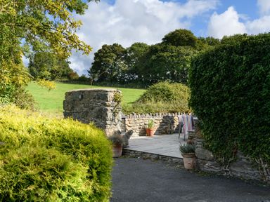 An outdoor patio area with stone walls potted plants and chairs in a garden with green bushes and trees at Ty Bach - Beaumaris in Beaumaris
