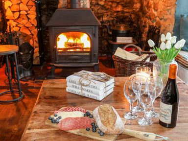 A wooden table with a loaf of bread sliced meat and cheese blueberries wine glasses a bottle of wine flowers and a candle in front of a wood burning stove at Ty Bach - Beaumaris in Beaumaris