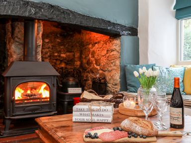 A fireplace with a burning wood stove and a wooden table with bread, salami, blueberries, wine bottle, glasses, flowers, books, and candle at Ty Bach - Beaumaris in Beaumaris