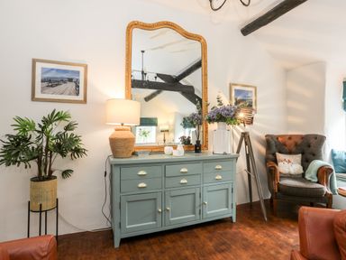 A room with a green cabinet topped with a large mirror and lamp a plant in a wicker basket and an armchair with pillows at Ty Bach - Beaumaris in Beaumaris