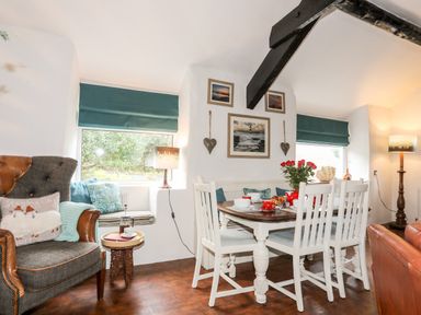 A dining area with a wooden table, white chairs, a high-backed armchair, windows with teal blinds, and framed pictures on the wall at Ty Bach - Beaumaris in Beaumaris