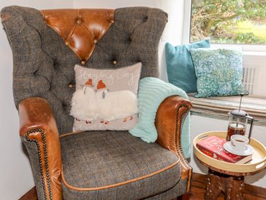 A brown and gray armchair with a pillow and blanket next to a window with cushions and a small round wooden table with a book a cup of coffee and a french press at Ty Bach - Beaumaris in Beaumaris