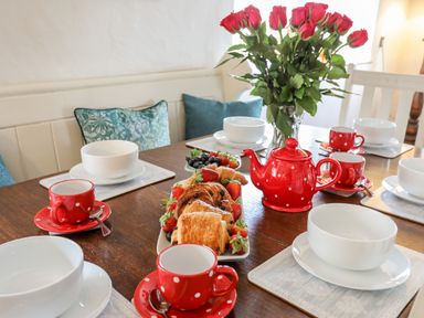 A dining table set with white bowls red polka dot cups and a red teapot with pastries and fruit and a vase of red roses at Ty Bach - Beaumaris Beaumaris
