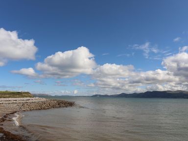 A coastal scene with a pebble beach and calm water under a blue sky with clouds at Ty Bach - Beaumaris in Beaumaris