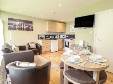 A small living area with a sofa, armchairs, a kitchen with wooden cabinets, a dining table set with plates and glasses, and a wall-mounted TV at 107 Kings Chalet Park in Cromer