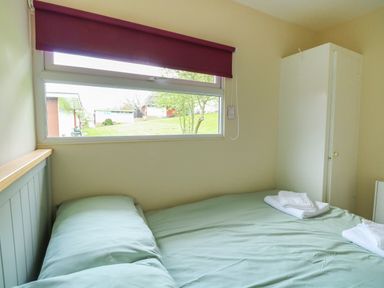 A bedroom with a bed covered in green bedding, two folded white towels on the bed, a white wardrobe, and a window with a maroon blind at 107 Kings Chalet Park in Cromer