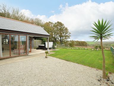An outdoor area with a gravel patio, grassy lawn, small palm tree, wooden building with large windows, hot tub and metal garden table at Pol Glas Cabin in Churchtown near Mullion