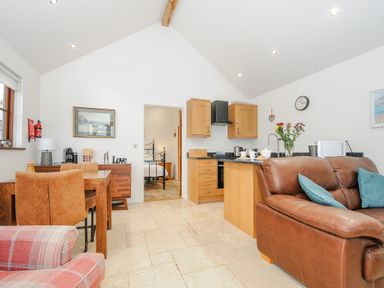 A living area with a brown leather sofa, wooden dining table with chairs, and a kitchenette at Pol Glas Cabin in Churchtown near Mullion