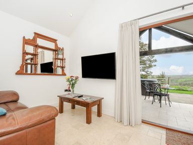 A living room with a brown leather sofa a wooden coffee table a wall-mounted tv and a large mirror next to glass doors leading to an outdoor seating area with a view at Pol Glas Cabin in Churchtown near Mullion