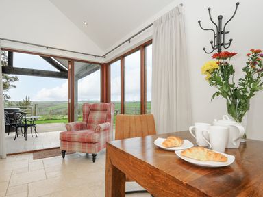 A dining table with croissants cups and teapot next to armchair and large windows at Pol Glas Cabin Churchtown near Mullion