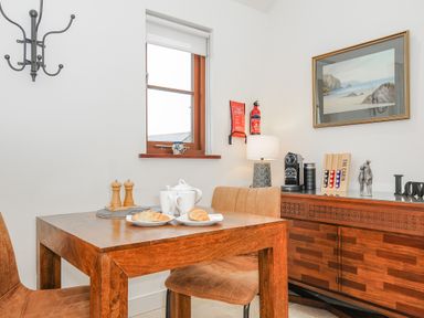 A wooden dining table with plates croissants and tea set next to a window and sideboard with coffee machine and lamp at Pol Glas Cabin in Churchtown near Mullion