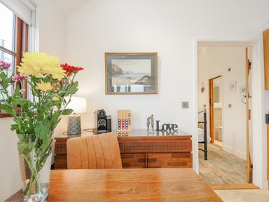 A dining area with a wooden table a chair flowers in a glass vase a sideboard with a coffee machine lamp and decorative items at Pol Glas Cabin in Churchtown near Mullion