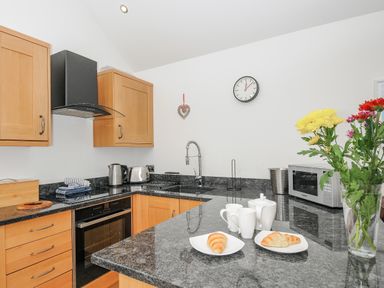 A kitchen with wooden cabinets black countertop a toaster kettle microwave plates with croissants and a vase of flowers at Pol Glas Cabin in Churchtown near Mullion