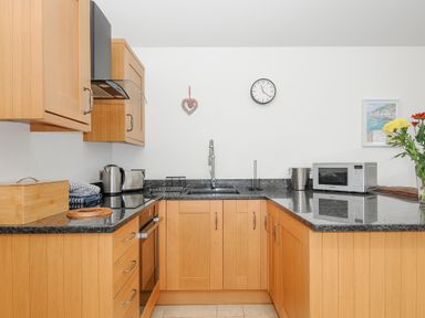 A kitchen with wooden cabinets and black countertops a microwave clock and flowers at Pol Glas Cabin in Churchtown near Mullion