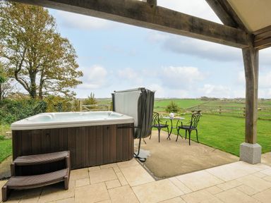 An outdoor patio with a hot tub small table with two chairs and a view of a green field at Pol Glas Cabin in Churchtown near Mullion