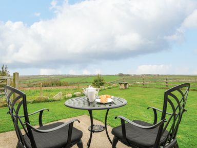 A garden with a metal table and two chairs with tea set and croissants overlooking fields at Pol Glas Cabin Churchtown near Mullion