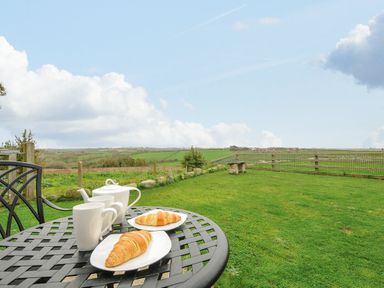 A garden with a metal table and chairs holding tea cups a teapot and croissants with fields and a fence in the background at Pol Glas Cabin in Churchtown near Mullion