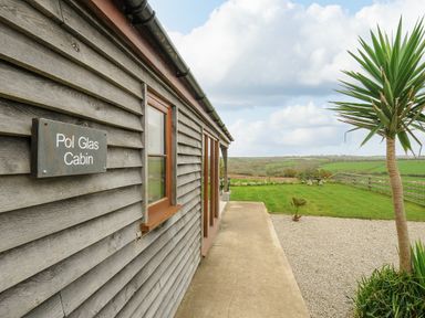 Exterior side view of a wooden cabin with a sign reading Pol Glas Cabin and a garden with grass trees and a gravel area at Pol Glas Cabin Churchtown near Mullion