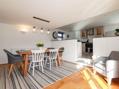 A dining area with a wooden table white and gray chairs a striped rug and a kitchen in the background at Great Meadow Barn in Crantock