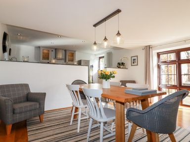 A dining area with a wooden table and mixed chairs next to a living area and kitchen at Great Meadow Barn in Crantock