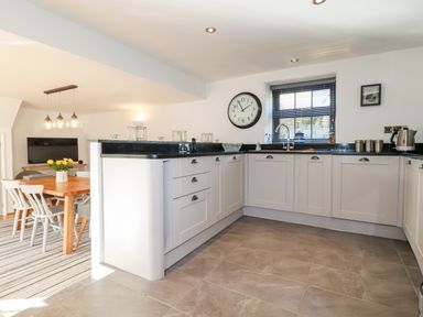 A kitchen with white cabinets a black countertop and a dining area with a wooden table and chairs at Great Meadow Barn in Crantock
