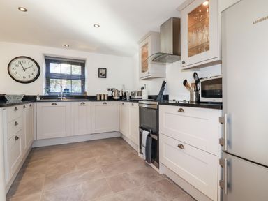 A kitchen with white cabinets black countertops a stainless steel oven and refrigerator a wall clock and a window with blinds at Great Meadow Barn in Crantock