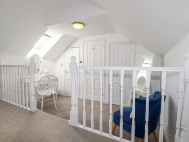 An attic landing area with a white desk and chair under a skylight window and a blue armchair with a mirror behind it at Great Meadow Barn in Crantock