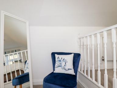 A blue chair with a sailboat design pillow next to a white railing and a mirror reflecting part of a bedroom at Great Meadow Barn in Crantock