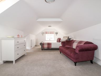 A bedroom with a white dresser, a bed with white and pink bedding, and a burgundy sofa at Great Meadow Barn in Crantock