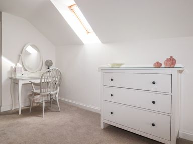 A room with a white dressing table and chair under a skylight and a white three-drawer dresser with decorative items at Great Meadow Barn in Crantock