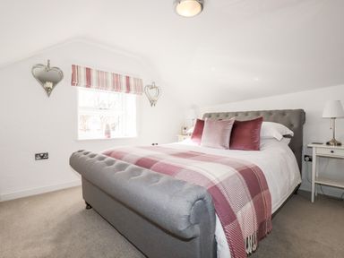 A bedroom with a grey upholstered bed with pink cushions and a pink checkered throw with white bedside tables and lamps at Great Meadow Barn in Crantock