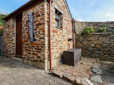 An outdoor area with a stone building and a wooden door a plastic storage box and a stone wall with plants at Great Meadow Barn in Crantock