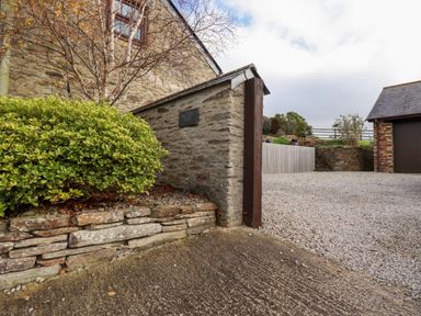 A stone wall with a green bush beside a gravel driveway and a building with a dark door at Great Meadow Barn in Crantock